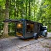 SylvanSport VAST travel trailer parked in a forest setting with open side and outdoor kitchen.