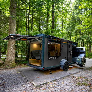 SylvanSport VAST travel trailer parked in a forest setting with open side and outdoor kitchen.