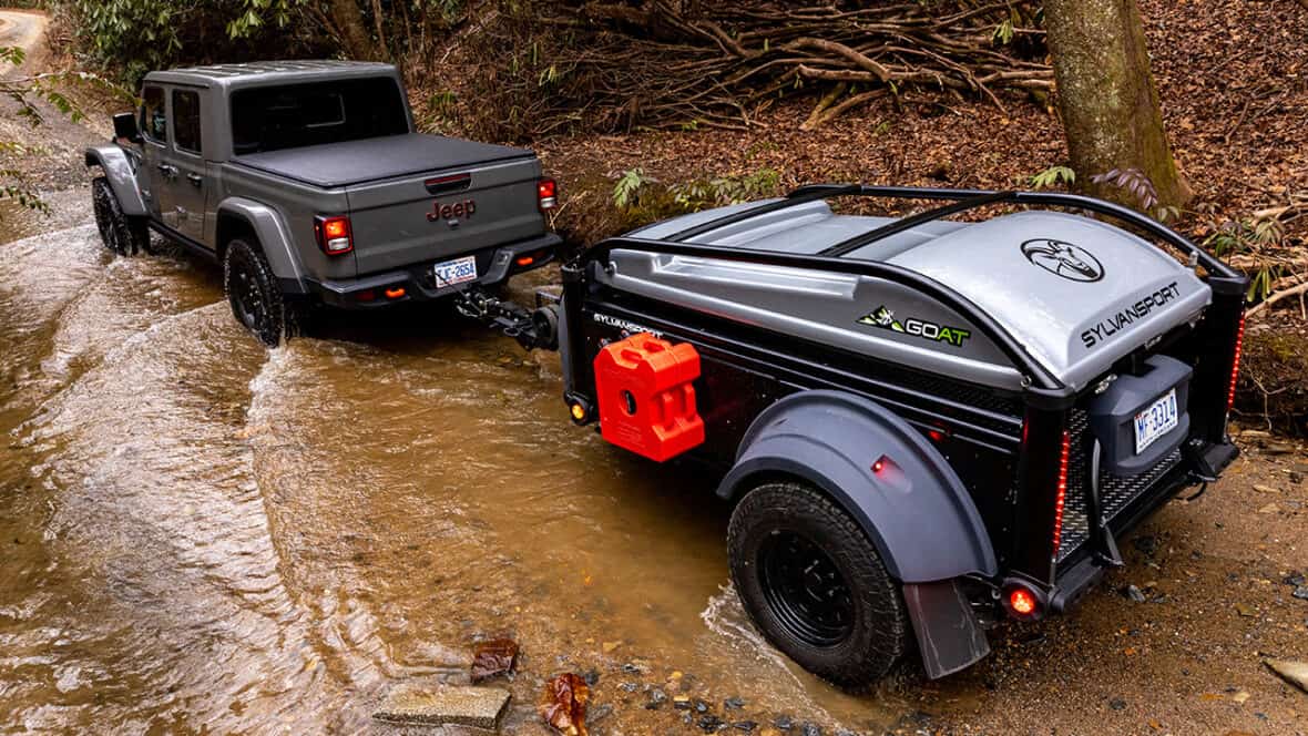 Off-road camper trailer attached to a Jeep in a forest stream.