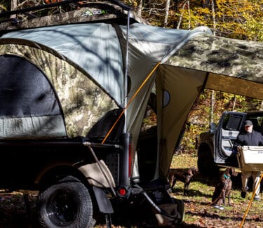 A rugged SylvanSport camping trailer with a pop-up tent in a forest setting.