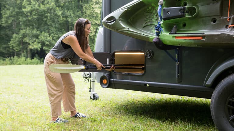 Woman loading a grill into a SylvanSport VIRE travel trailer on a grassy field.