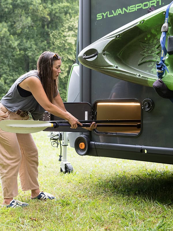 Woman loading a kayak into a SylvanSport VIRE travel trailer at a campsite.